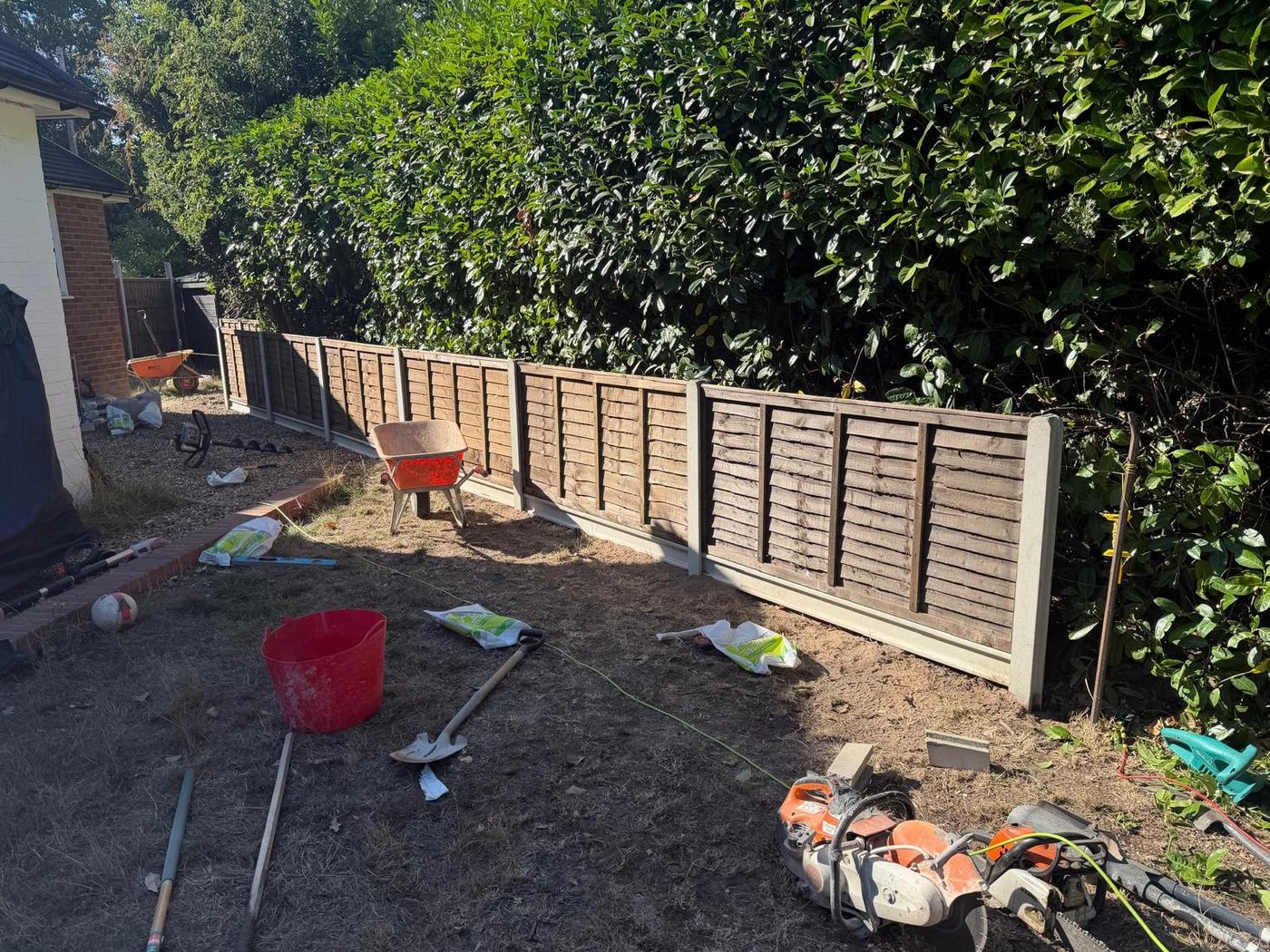 Lap fence panels on concrete posts between a bungalow and a laurel hedge with tools laid out