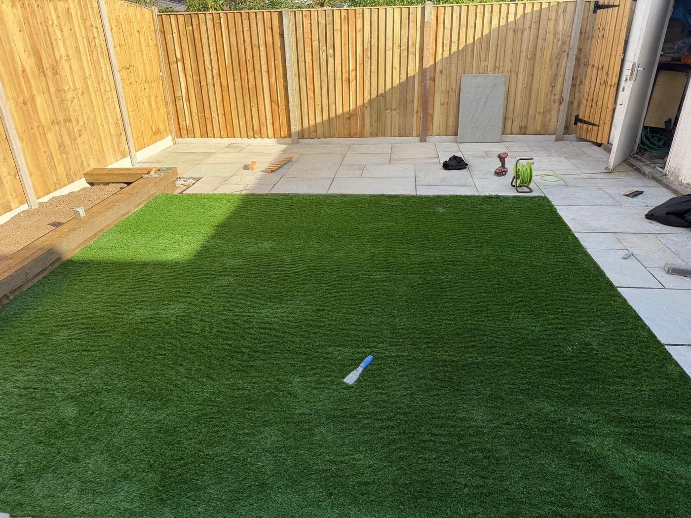 Artificial lawn bordered by grey stone paving inside a new closeboard fence on concrete posts