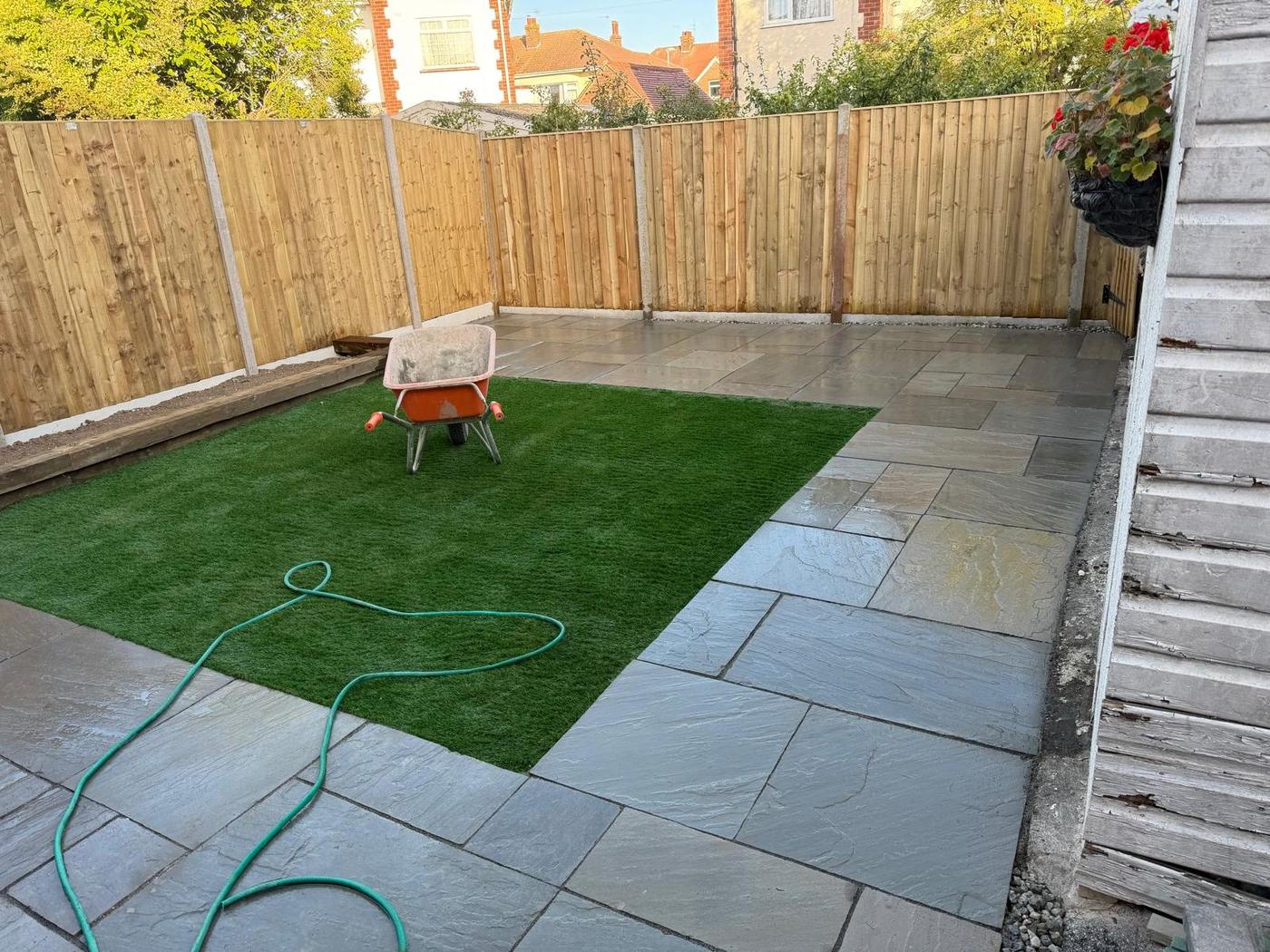Grey stone paving wrapping two sides of an artificial lawn, closeboard fencing behind