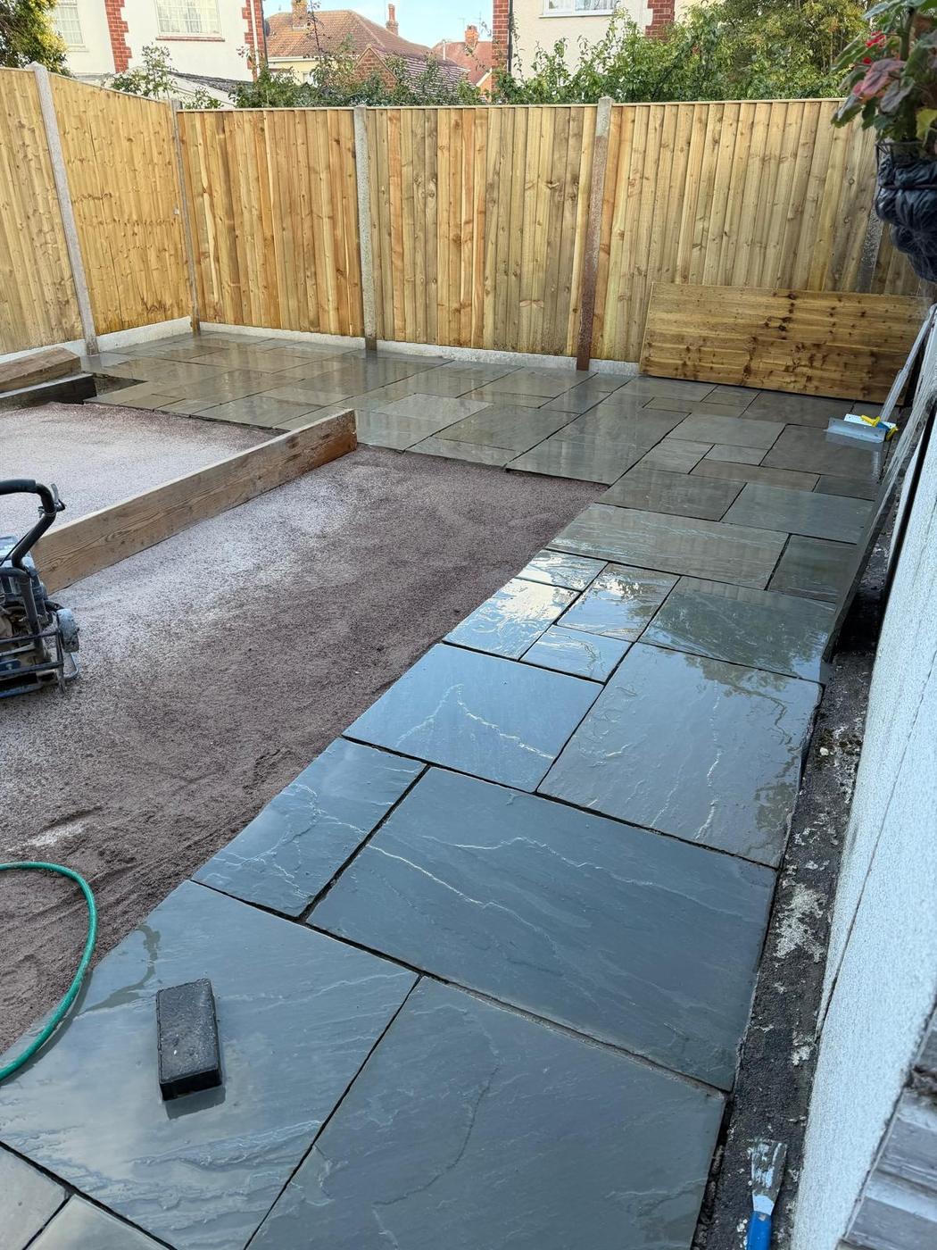 Grey paving slabs being laid on a mortar bed beside a new closeboard fence